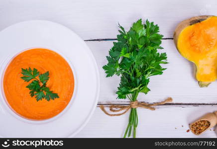 creamy pumpkin soup in a round white plate and a bunch of green parsley on a white wooden background, top view