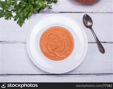 creamy pumpkin soup in a round white plate and an iron spoon on a white wooden table, top view