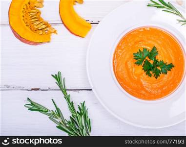 creamy pumpkin soup in a round white plate and pieces of fresh pumpkin on a white table, top view
