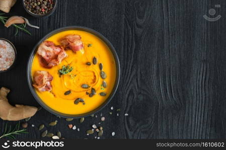 Creamy pumpkin soup with sauteed bacon slices with cream and pumpkin seeds in a black bowl on a black wooden table. Top view with copy space. Ingredients for making pumpkin soup on the table