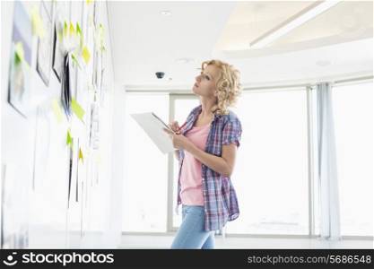 Creative businesswoman looking at papers stuck on wall while writing notes in office