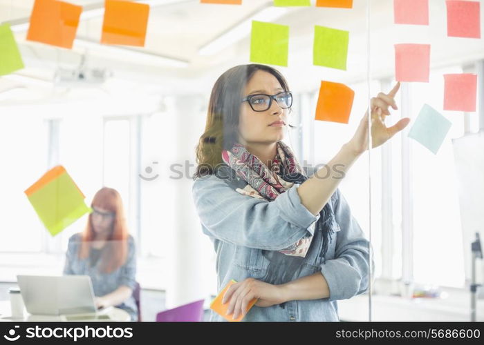 Creative businesswoman reading sticky notes on glass wall with colleague working in background at office