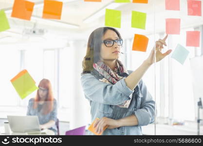 Creative businesswoman reading sticky notes on glass wall with colleague working in background at office