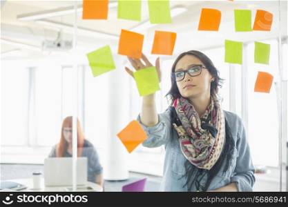 Creative businesswoman reading sticky notes on glass wall with colleague working in background at office