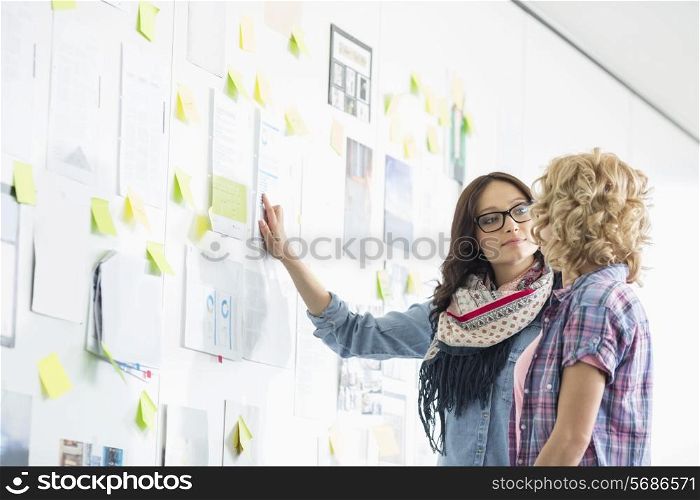 Creative businesswomen discussing over papers stuck on wall in office
