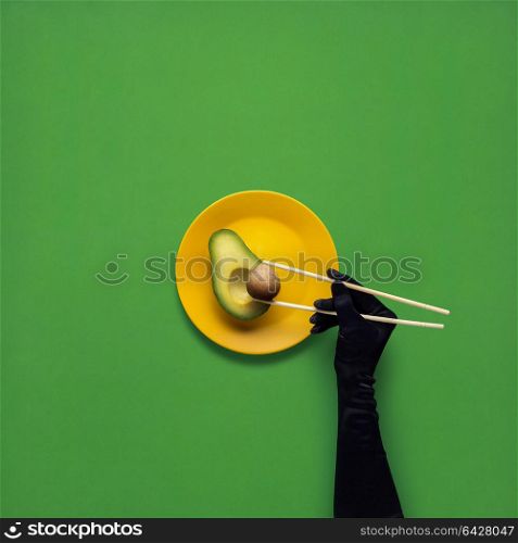 Creative concept photo of avocado with hand and chopsticks on painted plate on green background.