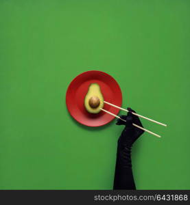 Creative concept photo of avocado with hand and chopsticks on painted plate on green background.