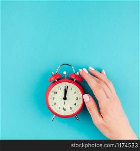 Creative flat lay concept top view of woman hand holding the red vintage alarm clock on square bright blue turquoise color paper background with copy space in minimal style, template for text