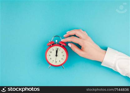 Creative flat lay concept top view of woman hand in white shirt holding the red vintage alarm clock on bright blue turquoise color paper background with copy space in minimal style, template for text