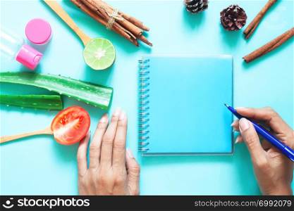 Creative flat lay of skincare and healthcare concept. Woman&rsquo;s hands with empty notebook, tomato, aloe vera, lime and cosmetic containers on pastel background
