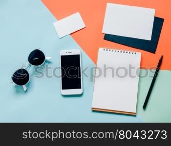 Creative flat lay of workspace desk with smartphone, blank notebook, envelope, name card and sunglasses on minimal color background