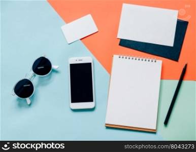 Creative flat lay of workspace desk with smartphone, blank notebook, envelope, name card and sunglasses on minimal color background