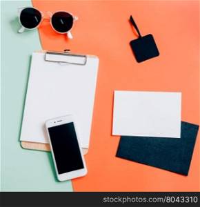 Creative flat lay of workspace desk with smartphone, clipboard, envelope, tag and sunglasses on minimal color background