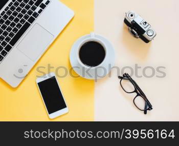Creative flat lay photo of workspace desk with laptop, smartphone, coffee, eyeglasses and film camera on yellow modern background&#xA;