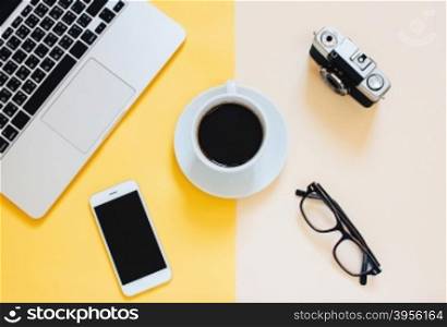 Creative flat lay photo of workspace desk with laptop, smartphone, coffee, eyeglasses and film camera on yellow modern background&#xA;