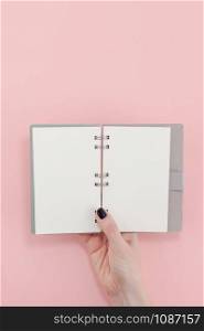Creative flat lay top view of woman hands black manicure with empty notebook mock up on pastel millennial pink paper background copy space. Minimal writing concept template for text lettering blogging