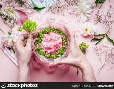 Creative Florist workspace. Female hands making pretty floral decoration arrangement with pink roses and green plant leaves in glass vase with water and florist decoration equipment, top view