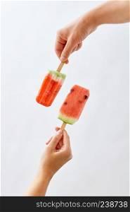Creative food composition of a fruit ice cream in a female hand and a juicy piece of watermelon on a stick holds a male hand on a gray background space for text. Summer dessert. A man&rsquo;s hand holds a slice of watermelon on a stick, in the form of an ice cream, and a red ice cream lolly in a woman&rsquo;s hand on a gray background with a copy of the space.