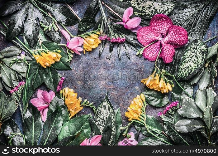 Creative frame made of various tropical flowers and leaves on dark rustic background, top view
