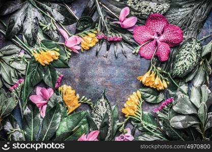 Creative frame made of various tropical flowers and leaves on dark rustic background, top view