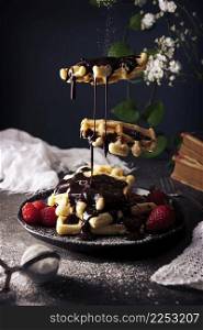 Creative image of homemade waffles levitating with liquid chocolate, fruits and powdered sugar on dark dish. Darkfood Levitation Photo.