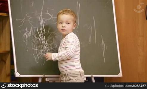 Creative little boy drawing on a chalkboard in a kindergarten classroom turning to look back over his shoulder for approval