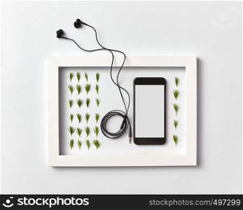 Creative ogranic herbal pattern of pine needles twigs, mobile smartphone with blank screen for mockup and headphones in a frame on a light gray background. Place for text. Flat lay.. Frame with organic pine needles pattern, smartphone mock-up and headphones on a light background.