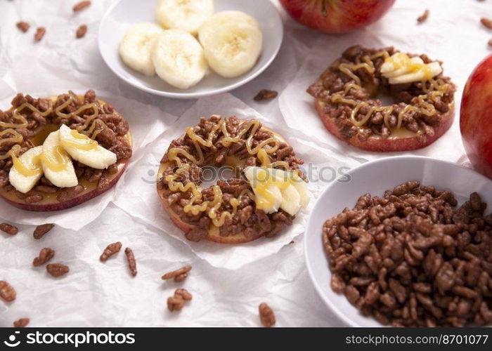 Creative party snack for holidays. Apple rounds with peanut butter, caramel and chocolate flavor puffed rice topping with banana slices. Funny appetizer for kids and adults.