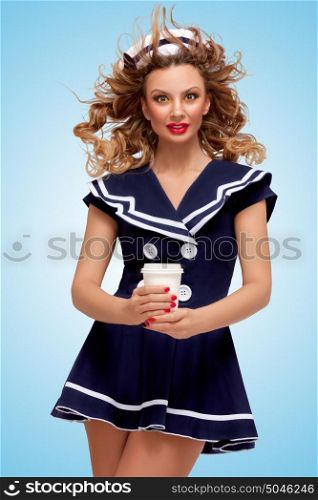 Creative photo of a playful pin-up sailor girl with a coffee-to-go cup, having wind in her hair on blue background.