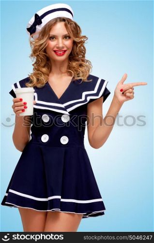 Creative photo of a playful pin-up sailor girl with a coffee-to-go cup, pointing aside with a finger on blue background.