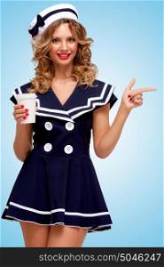 Creative photo of a playful pin-up sailor girl with a coffee-to-go cup, pointing aside with a finger on blue background.