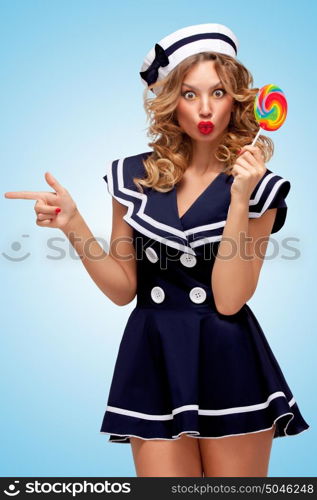 Creative photo of a playful pin-up sailor girl with a colorful lollipop, pointing aside with a finger on blue background.