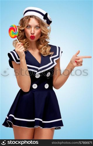 Creative photo of a playful pin-up sailor girl with a colorful lollipop, pointing aside with a finger on blue background.