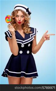 Creative photo of a playful pin-up sailor girl with a colorful lollipop, pointing aside with a finger on blue background.