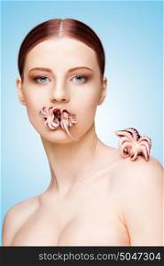 Creative portrait of a nude girl with beautiful face and body holding cooked baby octopus in mouth on blue background.
