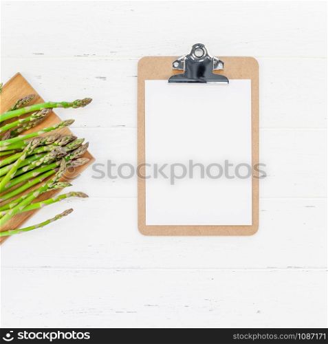 Creative scandinavian style flat lay top view mockup of fresh green asparagus paper clipboard on white wooden table background copy space. Minimal house cooking concept mock up for blog or recipe book