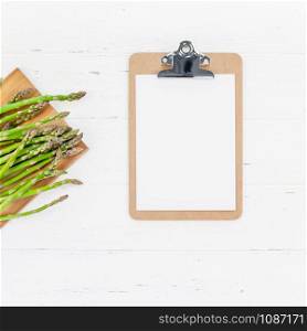 Creative scandinavian style flat lay top view mockup of fresh green asparagus paper clipboard on white wooden table background copy space. Minimal house cooking concept mock up for blog or recipe book