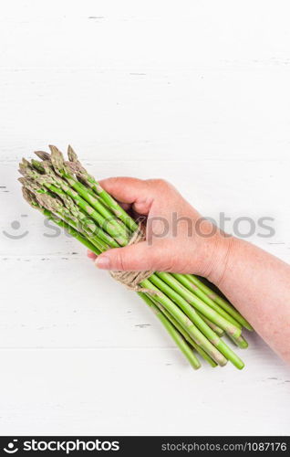 Creative scandinavian style flat lay top view of fresh green asparagus in elderly woman hands on white wooden table background copy space. Minimal house cooking concept for blog or recipe book