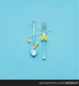 Creative still life photo of fork and spoon with raw pasta on blue background.