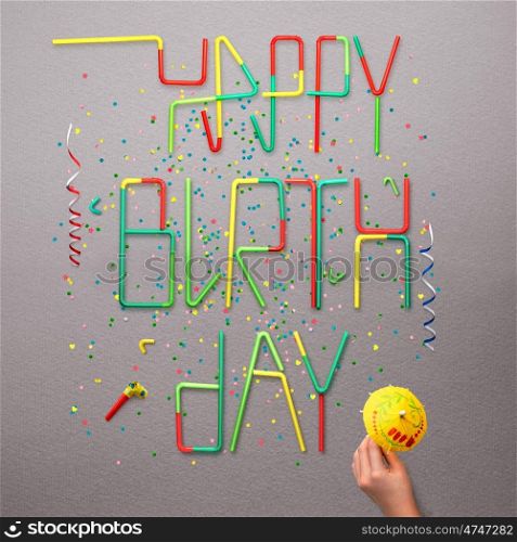 Creative still life photo of happy birthday sign made of cocktail straws with confetti and serpentine on grey background.