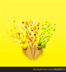 Creative still life photo of three waffle cones with candies, fruits and marshmallow on jellow background.