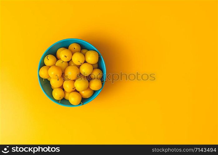 Creative summer pattern made of fresh yellow cherry plums in blue bowl on pastel yellow background. Fruit minimal concept. Flat lay.