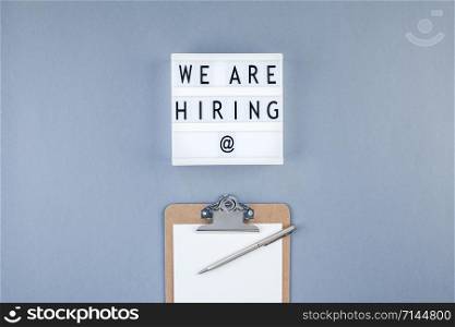 Creative top view flat lay of desk with we are hiring text on lightbox with copy space on blue background in minimal style. Concept of new job, hiring recruitment process, new team members screening