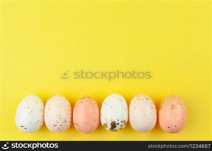 Creatively painted eggs in pastel colors decorated with gold leaf are arranged in row along bottom edge on yellow background, copy space. Happy Easter DIY concept. Flat lay. Horizontal.