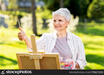 creativity, art and hobby concept - senior woman with easel and color palette painting outdoors. senior woman with easel painting outdoors