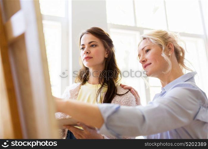 creativity, education and people concept - artists or student girl with palette and teacher discussing painting on easel at art school studio. artists discussing painting on easel at art school