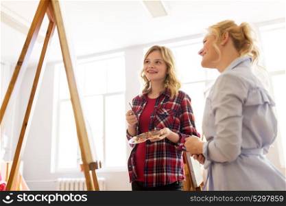 creativity, education and people concept - artists or student girl with palette and painting knife and teacher discussing picture on easel at art school studio. artists discussing painting on easel at art school