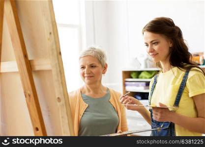 creativity, education and people concept - female artists or student girl with brush and palette and teacher painting on easel at art school studio. women with brushes painting at art school