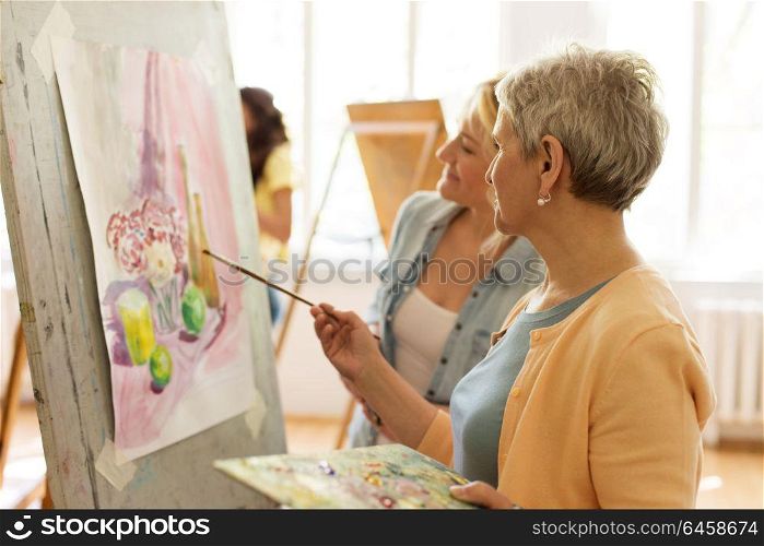 creativity, education and people concept - female artists or students with brushes and palettes painting on easel at art school studio. women with brushes painting at art school
