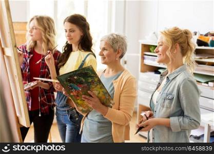 creativity, education and people concept - group of female artists or students with brushes and palettes and teacher painting on easel at art school studio. women with brushes painting at art school